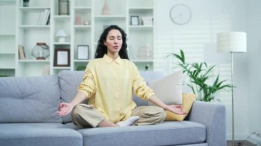 Genç esmer kadın, evde oturma odasında nilüfer çiçekli koltukta oturmuş, gözleri kapalı meditasyon yapıyor. Sakin, mutlu esmer bir kadın yoga yapar, derin derin nefes alır, rahat bir kanepede dinlenir.