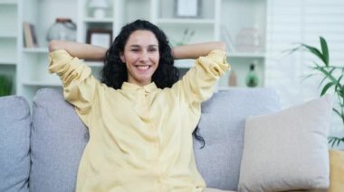 Genç gülümseyen kıvırcık esmer kadın oturma odasında otururken gözleri kapalı ve elleri başının arkasında dinleniyor. İşten sonra evdeki kanepede dinlenen gömlekli mutlu bir kadın.