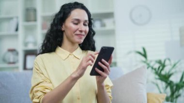 Evdeki oturma odasında kanepede otururken akıllı telefon kullanan mutlu genç bir kadın. Gülümseyen kadın sosyal ağlara takılır, çevrimiçi bir mağazadan alışveriş yapar, arkadaşlarıyla sohbet eder, mesajlar yazar.