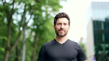 Siyah tişörtlü, kameraya bakan mutlu sakallı yakışıklı adamın portresi. Yetişkin bir sporcu, şehir dışında şehir arka planında koşuyor. Şehir caddesinde sağlıklı bir yaşam tarzı sabah sporu yapıyor.