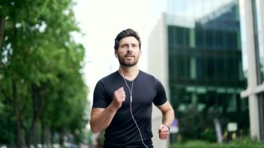 Yakışıklı sakallı bir erkek koşucu şehrin modern bir arka planında koşuyor. Kulaklıkla koşan sporcu adam dışarıda koşuyor. Şuna bak. Bir fitness bilekliği ya da akıllı yama.