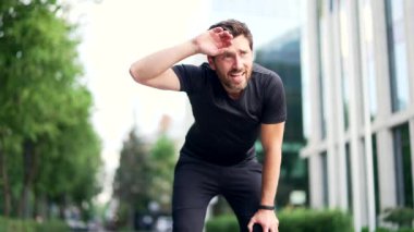 Şehir caddesinde koştuktan sonra yorgun koşucunun nefes darlığı var. Yorgunluktan bitkin düşmüş koşan yetişkin erkek elleri dizlerinin üstünde duruyor, derin derin nefes alıyor, alnındaki teri siliyor.