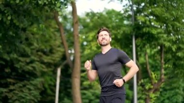 Yakışıklı sakallı erkek koşucu şehir parkında koşuyor sporcu atlet koşucusu dışarıda koşuyor. Şuna bakın, spor bilekliği ya da akıllı erkek temiz havada sabah aktivitelerinden hoşlanıyor.