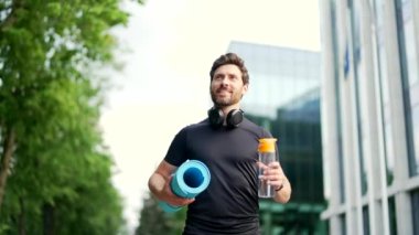 Sakallı, yakışıklı, spor salonuna ya da elinde bir şişe su ve minderle yoga antrenmanına giden mutlu bir adam. Parkın dışına doğru yürüyün. Fitness erkeği golün tadını çıkarın.