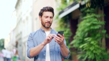 Neşeli yetişkin adam zaferi kutluyor telefonda harika haberler okuyor. Tatmin olmuş erkek, dışarıdaki bir caddede dururken akıllı telefona bakarak olumlu bir mesaj alır.