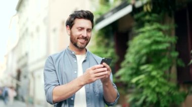 Mutlu, sakallı adam akıllı telefon uygulamasını şehir caddesinde kullanıyor. Açık havada mesajlaşan, sohbet eden cep telefonlarına bakıyorum. Erkek, sosyal medyayı karıştırıyor. Gülümsüyor. teknoloji ve yaşam tarzı kavramı