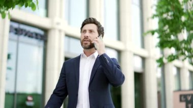 Kendine güvenen, sakallı, yetişkin, resmi takım elbiseli bir iş adamı. Şehir caddesinde ofis binasının önünde cep telefonuyla konuşuyor. İş adamı, gülümseyen mutlu girişimci iletişim kurar.