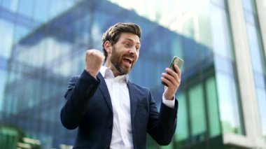 Neşeli iş adamı mutlu bir okuyucu cep telefonundan harika haberler alıyor. Resmi takım elbiseli bir iş adamı, şehrin caddesindeki ofis binasının yanında duran akıllı telefona bakarak zaferi kutluyor.