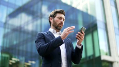 Yetişkin iş adamı, dışarıdaki şehir caddesindeki modern iş merkezinin yanında duran akıllı telefon ekranına bakarak kötü haber mesajı alıyor. Sinirli yönetici