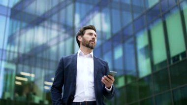 Kendine güvenen, takım elbiseli, olgun sakallı bir iş adamı modern cam ofis binasının yakınında elinde akıllı telefon tutarak yürüyor. Erkek girişimci, yatırımcı, seo ya da yönetici dışarıda cep telefonu kullanıyor