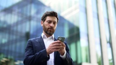 Cep telefonu kullanan yakışıklı sakallı bir iş adamı. Yetişkin bir adam mesaj atıyor, daktilo kullanıyor, akıllı telefon kullanıyor şehir merkezindeki iş merkezinin yanında duruyor.