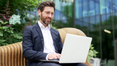 Takım elbiseli yakışıklı bir iş adamı laptopuyla modern ofis iş merkezinin yanındaki bankta oturuyor Mutlu Çalışanların Yöneticisi dışarıdaki caddede bilgisayarlara göz gezdirip gülümsüyor.