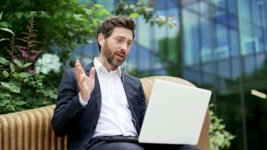İş adamı, binanın dışındaki modern bir ofis binasının yanındaki bankta otururken uzaktan iletişim kuruyor. Takım elbiseli iş adamı sakallı bir girişimci internette konuşuyor. Açık hava.