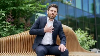 Aşırı çalışan olgun işadamı, modern iş merkezinin yanındaki bankta otururken kalp krizinde ani bir ağrı yaşıyor. Stresli erkek, kalp ağrısı çekiyor.