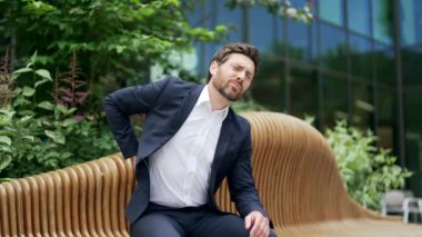 Yorgun girişimci işadamı ciddi bir sırt ağrısı çekiyor modern iş merkezinin yanındaki bankta oturuyor. Sağlıksız, hasta, sırtına kramp giren bir adam. Açık havada omurga ağrısı çekiyor.