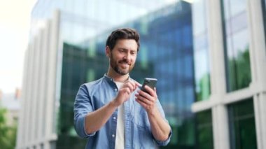 Modern iş yeri binasının yanında rahat duran yakışıklı, mutlu sakallı iş adamı elinde akıllı bir telefon, elinde olgun yetişkin erkek girişimci seo yöneticisi, dışarıda yaşam tarzı dışında bir cep telefonu kullanıyor.