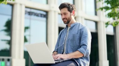 Modern ofis merkezinin yanındaki bankta oturan yakışıklı, sakallı iş adamı. Şehir sokaklarındaki gülüşünde dizüstü bilgisayarında geziniyor.