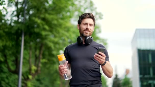 健康なスポーティな自信を持って成熟した大人のひげ付きハンサムな男は トレーニングや屋外でのトレーニングの後 市街地や公園を歩いています 携帯電話のスマートフォンを使用して 水のボトルを保持すると笑顔になります — ストック動画