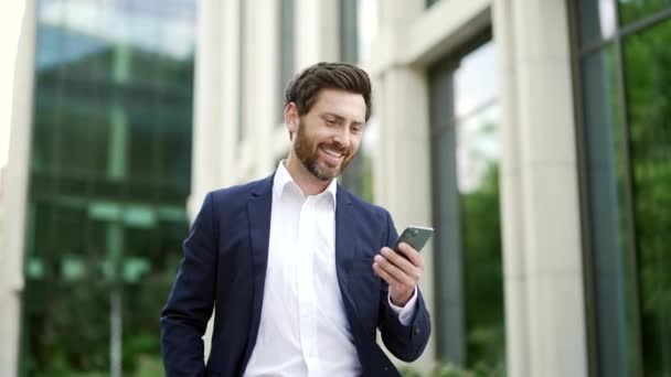 スマートフォンを手に持っている近代的なビジネスオフィスビルの近くを歩いてスーツに成熟した成熟したビジネスマン 起業家 投資家 ソウまたはマネージャーが外で携帯電話を読み取る — ストック動画