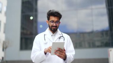 Bir hastane binasının önünde dururken beyaz önlüklü gülümseyen genç doktor tablet kullanıyor. Gözlüklü doktor iş arkadaşıyla internette sohbet ediyor ya da haberlere bakıyor.