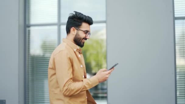 一个面带微笑的年轻人看着手机在街上走到绿松树旁的办公室 戴眼镜的英俊大胡子商人浏览智能手机 在网上聊天 — 图库视频影像