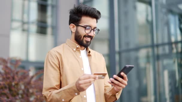一位面带微笑的年轻大胡子男子站在一幢办公楼外 用智能手机输入信用卡号码 戴眼镜的漂亮商人在网上购物 为好价钱感到高兴 — 图库视频影像