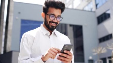 Kapatın. Genç, gülümseyen bir işadamı bir ofis binasının yanında caddede dururken akıllı telefon kullanıyor. Mutlu yakışıklı sakallı erkek e-postaları kontrol ediyor, internette sohbet ediyor, müşteriyle mesajlaşıyor.