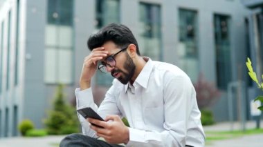 Beyaz gömlekli stresli genç adam, ofis binasının yanındaki bankta otururken akıllı telefondan kötü haberler okuyordu. Endişeli iş adamı üzgün, morali bozuk, telefondaki mesaja bakıyor.