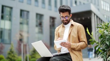 Gözlüklü yakışıklı öğrenci online webinar, sınıf web yayınlarını izlerken, dizüstü bilgisayara bakarken, e-öğrenirken, notlar alırken, video görüşmeleri yaparken, kampüsteki bir bankta otururken üniversite sınavlarına hazırlanırken.
