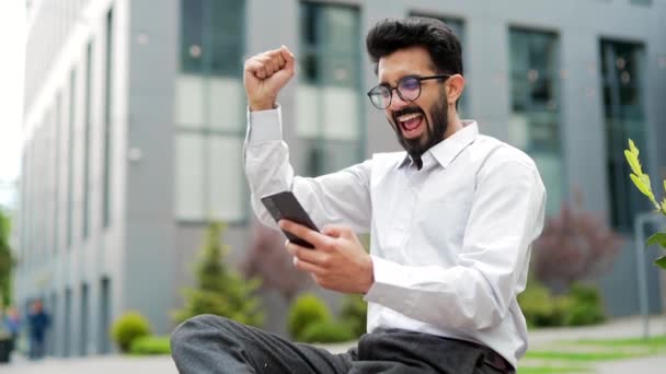 年轻的商人坐在办公大楼附近的一条长椅上 在智能手机上看到好消息 庆祝胜利 快乐的人牵着手做着胜利的手势 表示愿意 哇效果 — 图库视频影像