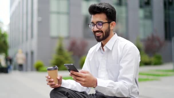 一位面带微笑的年轻商人坐在办公大楼附近的一条街上的长椅上 一边用智能手机 快乐英俊的大胡子男人检查电子邮件 网上聊天 发短信给客户 — 图库视频影像