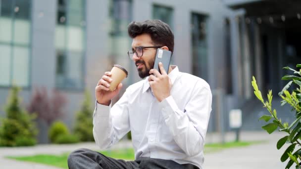 一位年轻的商人坐在办公大楼附近的一条街上的长椅上 一边用智能手机聊天 满脸胡须的自由撰稿人 与同事或客户交谈 喝咖啡 — 图库视频影像