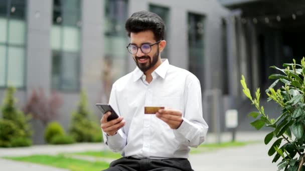 一位面带微笑的年轻商人站在一幢办公楼外 用智能手机输入信用卡号码 穿着白衬衫的帅哥在网上购物 为好价钱感到高兴 — 图库视频影像