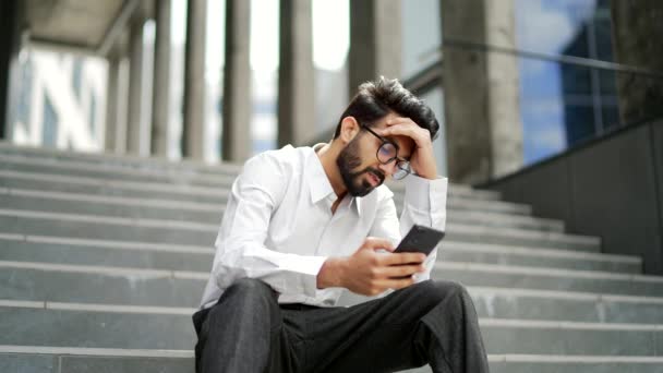 身穿白衬衫 心烦意乱的年轻人坐在一幢办公楼的楼梯上 一边用智能手机读坏消息 一边感到沮丧 忧心忡忡的商人看着电话里的留言 很伤心 心情不好 — 图库视频影像