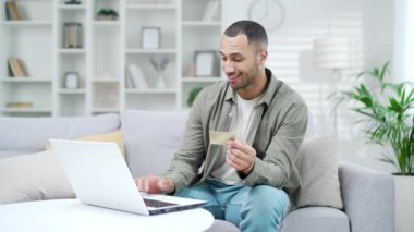 Adamın biri evdeki kanepede otururken kredi kartı ve dizüstü bilgisayarla ödeme yapıyor. İnternet mağazasından online alışveriş sipariş eden genç erkek müşteri e-bankacılık uygulaması için ödeme yapıyor