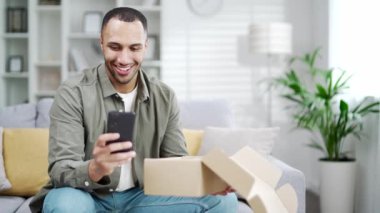 Mutlu genç adam internet mağazasından alışveriş yaparken paket siparişi aldı. Evde sosyal medyada olumlu geri bildirim için akıllı telefon kullanıldı. Tatmin olmuş erkek