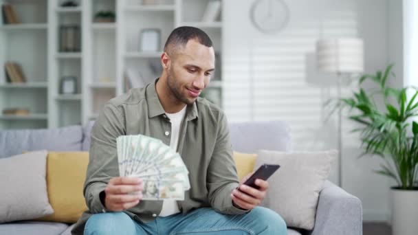 快乐的商人坐在家里挥动着一块钱的钞票扇子 微笑着拿着手机和钞票的男性投资者 看着相机 可以看到他们通过应用投资获得的盈利 — 图库视频影像