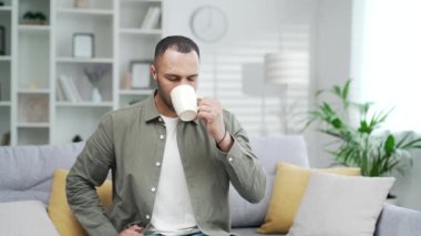 Genç ve mutlu bir adam bardaktan sıcak içecek içiyor. Memnuniyetle gülümseyen adam evdeki oturma odasında rahat koltukta çay yudumluyor. Kapalı mekanda karışık ırk erkekleri kahve ya da ilaç içerler.