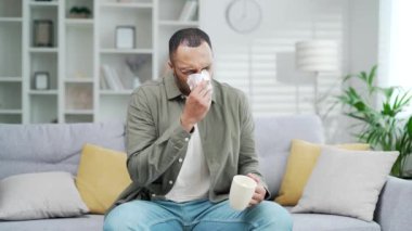 Sağlıksız hasta adam bardaktan sıcak içecek içiyor. Evdeki oturma odasındaki kanepede çay yudumlayan hasta bir adam. İç mekanda melez erkek soğuk algınlığı ya da grip kokusu bir içecek bardağı ya da ilaç.