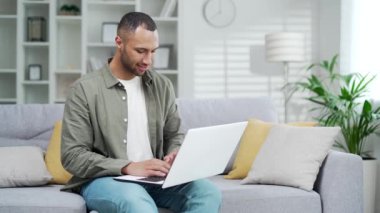 Genç ve yakışıklı Afrikalı Amerikalı adam evdeki kanepede otururken dizüstü bilgisayar kullanıyor. Karışık ırk erkek web 'de geziniyor ya da sosyal medya alışverişi yapıyor kapalı mekanda çalışma düzeniyle sohbet ediyor
