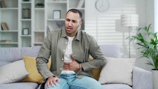 若い成人男性は 突然強い腹部胃痛 胃炎の問題を感じるソファに座っています 症状を有する男性は 自宅のアパートルームで下痢の消化管潰瘍膵炎を有する — ストック動画