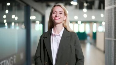Modern bir ofiste kollarını kavuşturmuş gülümseyen genç bir iş kadınının portresi. Şık, mutlu, sarışın, takım elbiseli girişimci kadın kameraya bakıyor. Başarılı menajer veya patronun vesikalığı