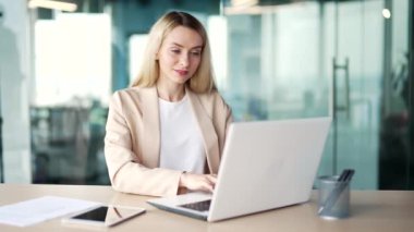 Modern bir ofisin çalışma masasında otururken dizüstü bilgisayarda yazan genç bir iş kadını. Bilgisayarda bir proje üzerinde çalışan sarışın kadın çalışan, bir müşteriye mesaj atan ya da internetten sohbet eden.