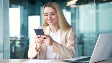 Takım elbiseli genç mutlu iş kadını modern ofiste iş yerinde otururken akıllı telefon kullanıyor. Gülümseyen sarışın kadın mesaj yazıyor, internette sohbet ediyor, sosyal medyada geziniyor ya da e-postaları kontrol ediyor.
