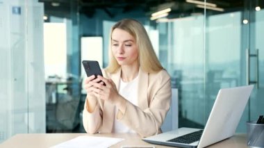 Takım elbiseli genç mutlu iş kadını modern ofiste iş yerinde otururken akıllı telefon kullanıyor. Gülümseyen sarışın kadın internetten alışveriş yapıyor, sosyal medyada geziniyor, mesaj yazıyor, müşteriyle sohbet ediyor.