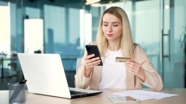 Modern ofiste masa başında otururken online alışveriş yapan genç bir iş kadını akıllı telefonuyla kredi kartı numarasını yazıyor. Gülümseyen ve tatmin olmuş sarışın bir kadın çalışan, mutlu bir şekilde alışveriş yapıyor.
