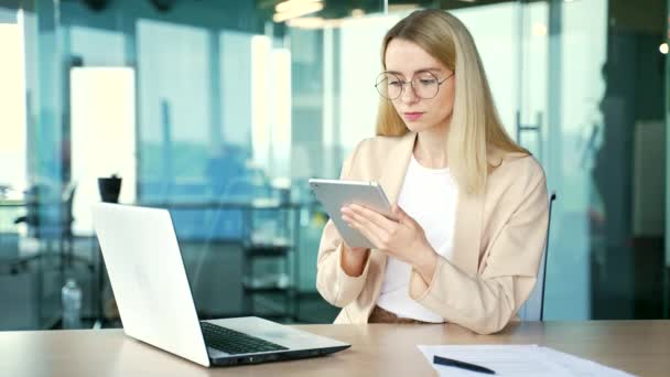 戴眼镜自信的年轻女商人坐在办公室工作时使用平板电脑 穿着西装的金发女员工在网上打字 浏览社交媒体 查看电子邮件 — 图库视频影像