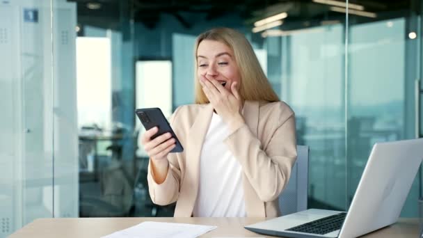 穿着西装的快乐而兴奋的年轻女商人坐在现代办公室的工作场所时 在智能手机上看到了好消息 女人庆祝胜利 摆出姿势 — 图库视频影像