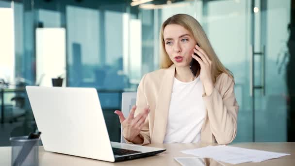 一位自信的女商人坐在现代办公室的办公桌前 一边用智能手机交谈 一个年轻的 面带微笑的金发女人与同事讨论生意或者与客户交谈 — 图库视频影像