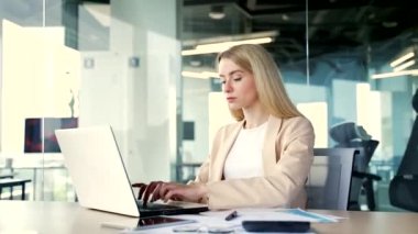 Yan görüş. Modern bir ofiste çalışırken dizüstü bilgisayarda yazan genç bir iş kadını. Bilgisayarda bir proje üzerinde çalışan sarışın kadın çalışan, bir müşteriye mesaj atan ya da internetten sohbet eden.